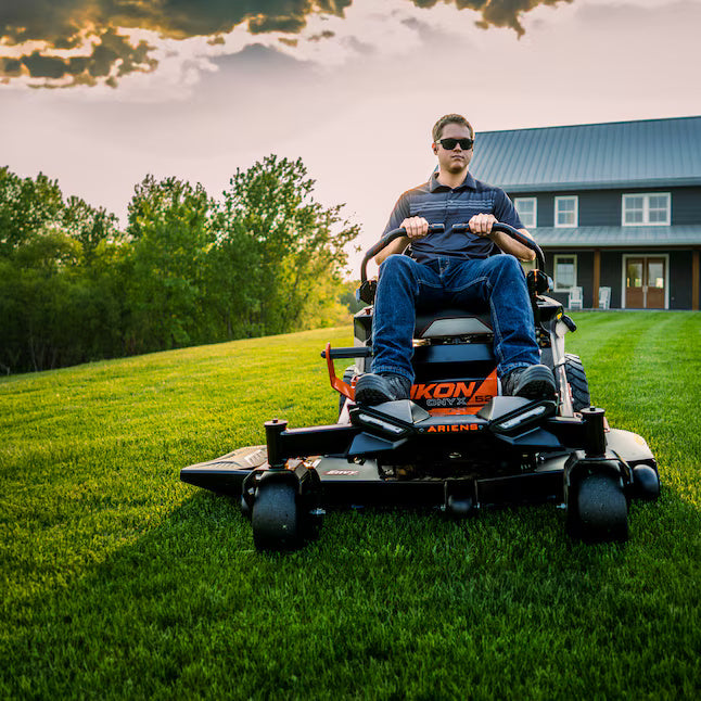 Man operating a lawn mower on a grassy field with a house and trees in the background