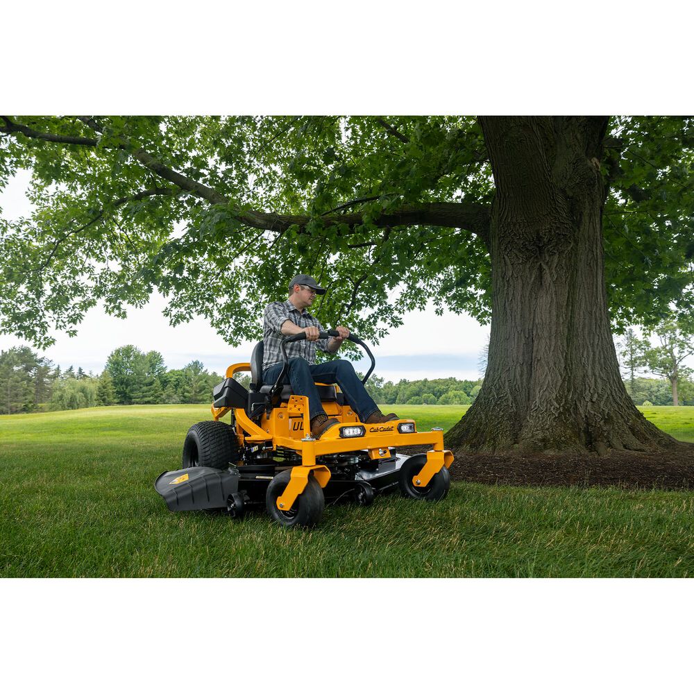 Man operating a yellow Cub Cadet lawn mower in a grassy area with trees.