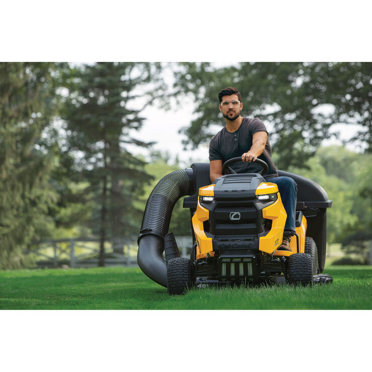 Man operating a yellow riding lawn mower in a grassy area with trees in the background