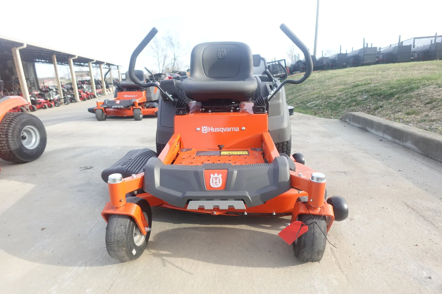 Orange and black Husqvarna riding lawn mower on a paved surface with grass and other equipment in the background.