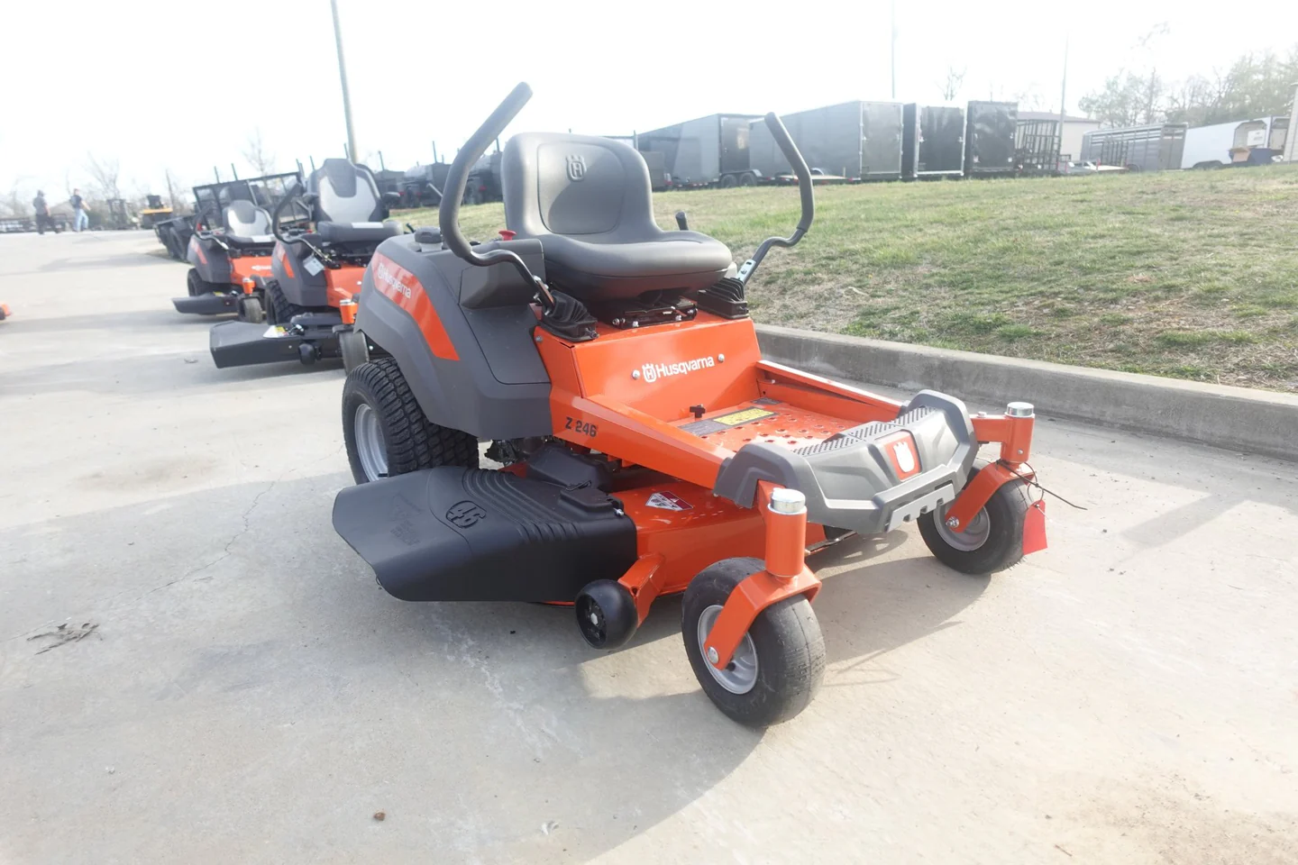 Orange and black riding lawn mower on a concrete surface with grass and vehicles in the background.