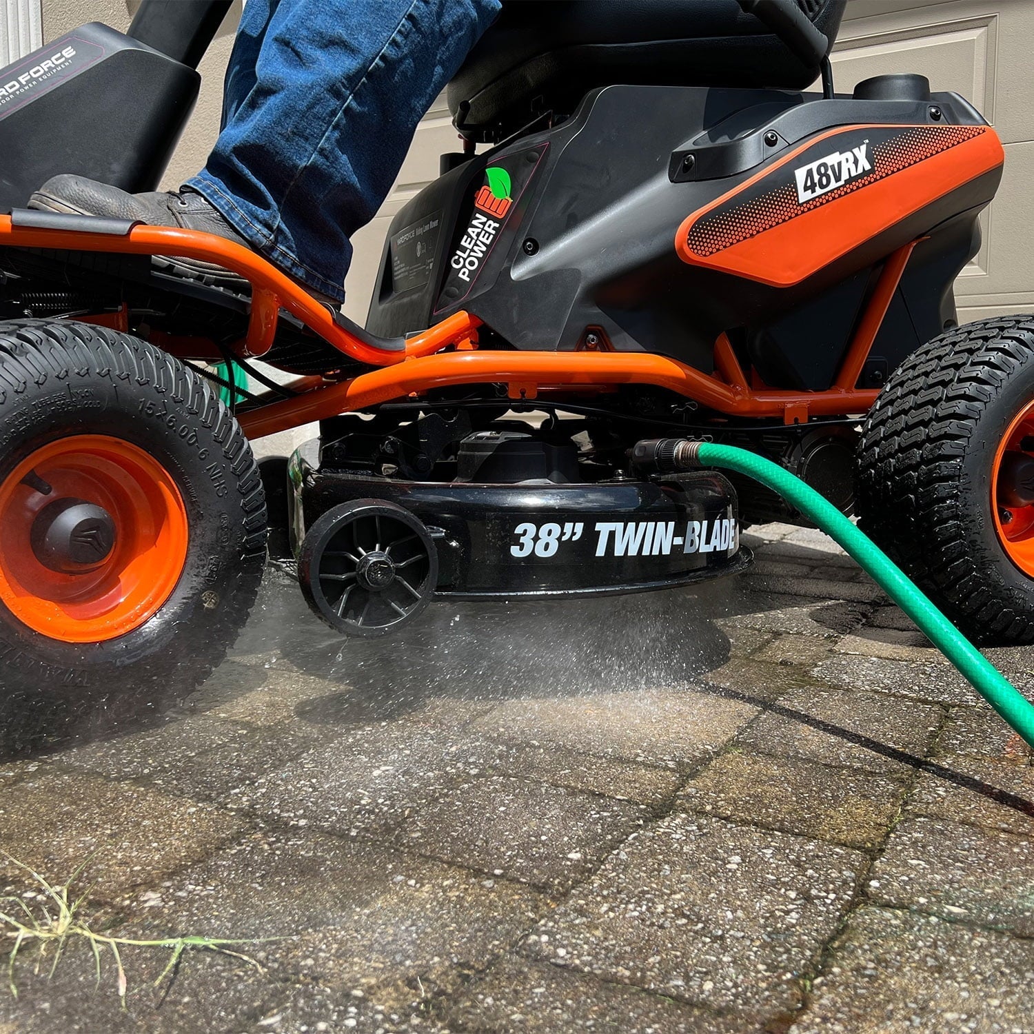 Person using a riding lawn mower with a hose attached on a driveway.