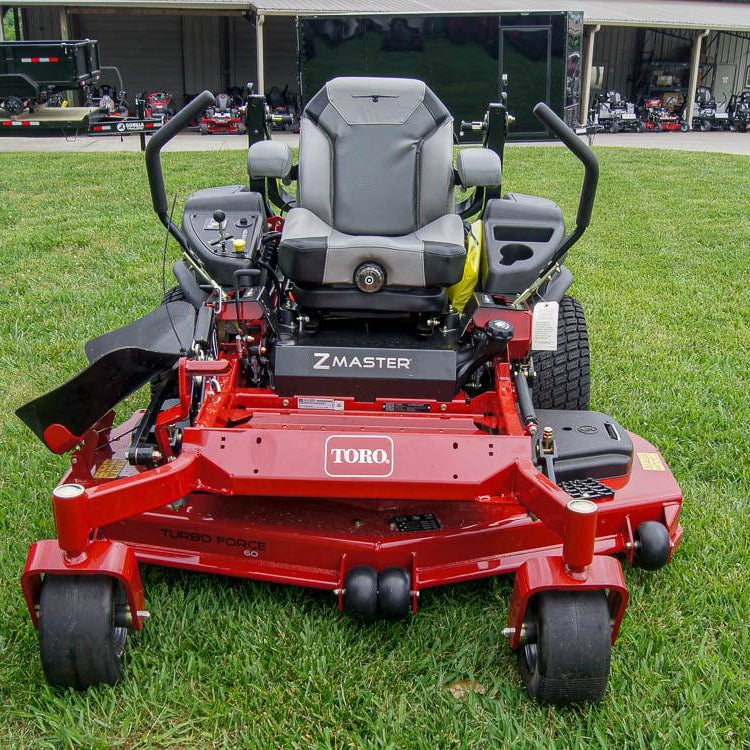 Red Toro zero-turn lawn mower on a grassy area with a building and vehicles in the background.