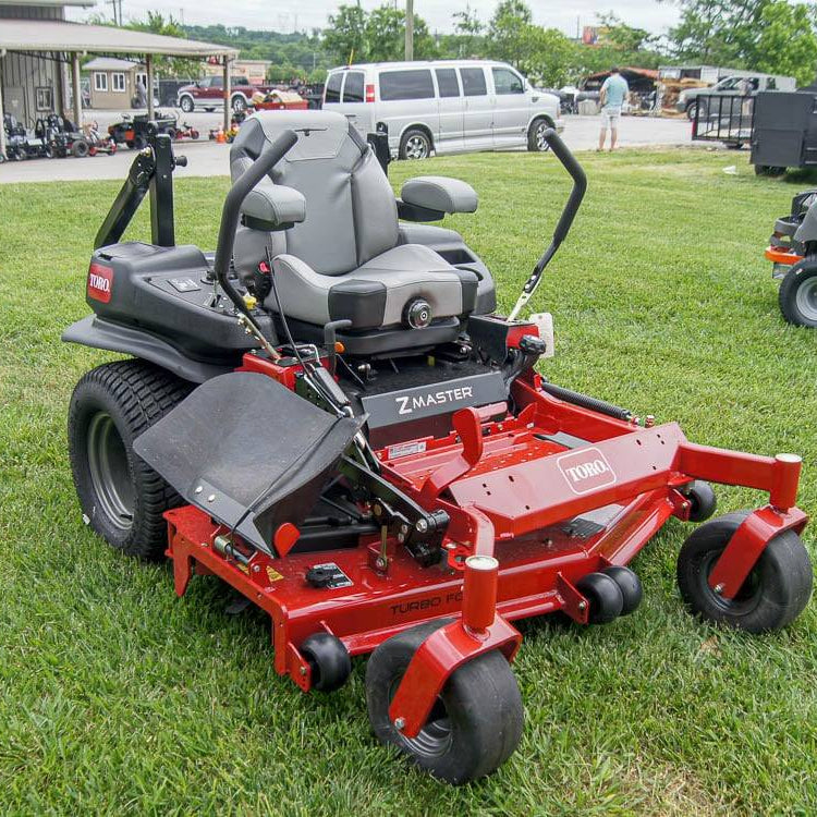Red zero-turn lawn mower on grass with a building and vehicles in the background